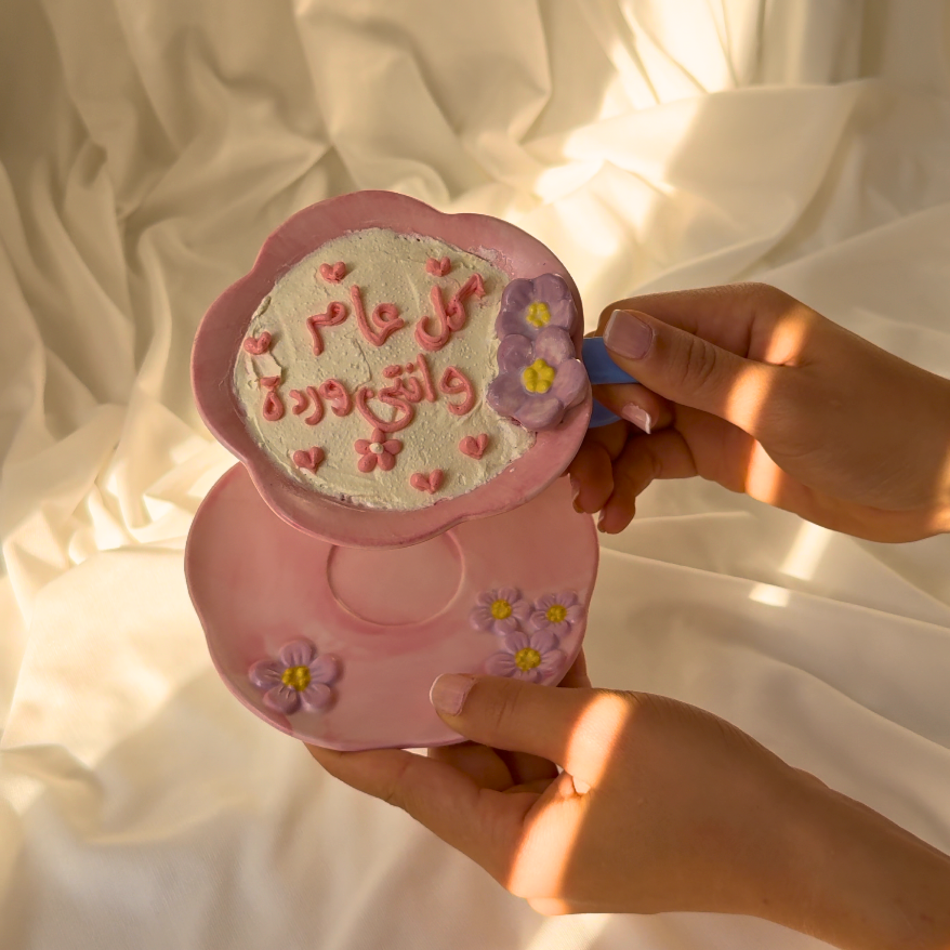 Pink decorative item with floral designs and text held by hands against a soft, light background