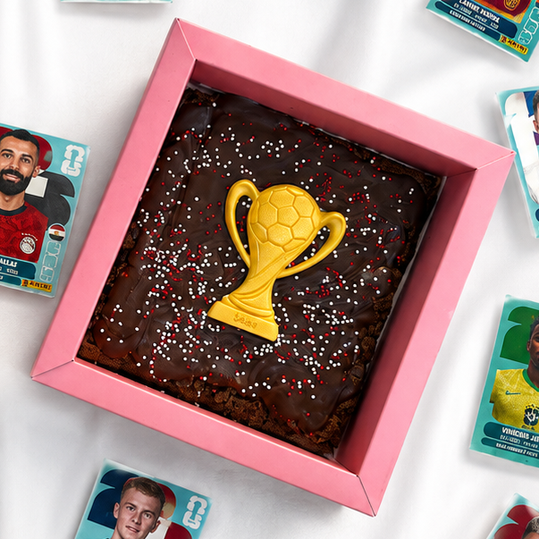 Chocolate brownie with a gold trophy on top in a pink box, surrounded by soccer-themed cards.