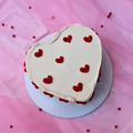 A heart-shaped cake decorated with white frosting and red hearts, presented on a pink background.