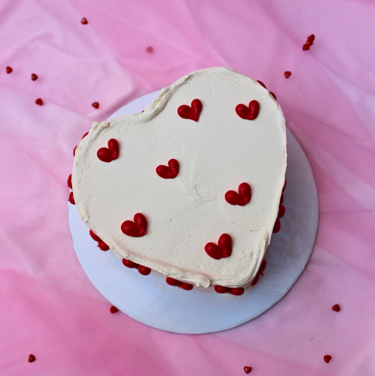 A heart-shaped cake decorated with white frosting and red hearts, presented on a pink background.
