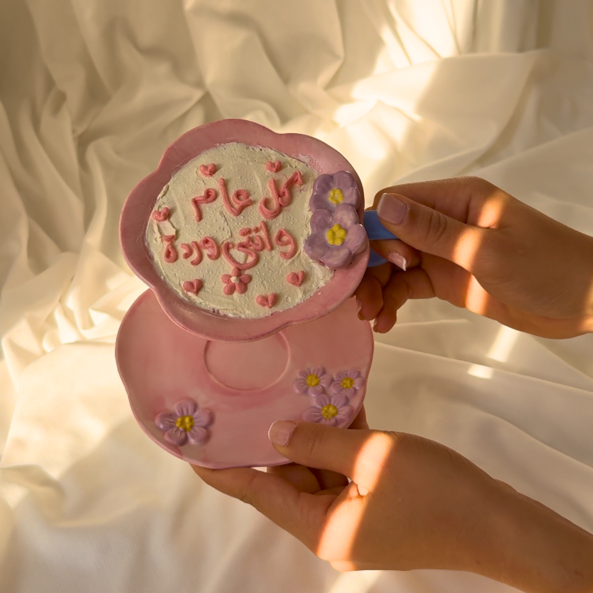 Pink decorative item with floral designs and text held by hands against a soft, light background