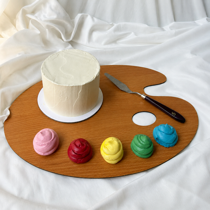 White cake on a wooden artist's palette with colorful frosting mounds and a knife.