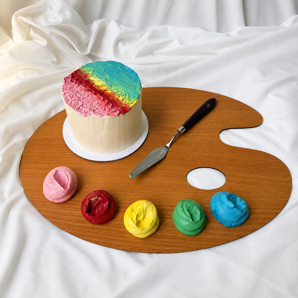 Colorful cake on a wooden artist's palette with frosting samples and a spatula.