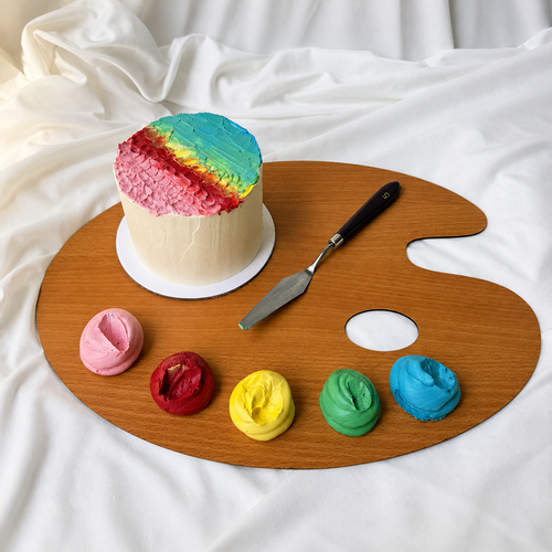 Colorful cake on a wooden artist's palette with frosting samples and a spatula.