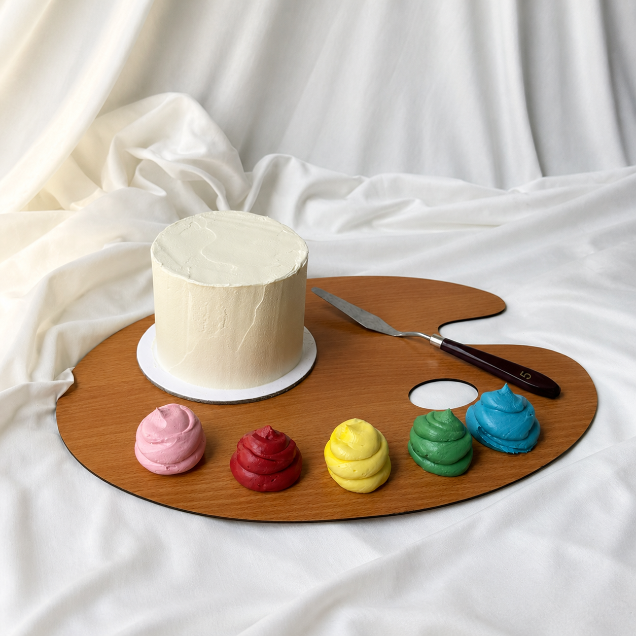 White cake on a wooden artist's palette with colorful frosting mounds and a knife on a white fabric background.
