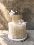 Two-tiered wedding cake with feathered bottom tier and floral decorations on a textured surface.
