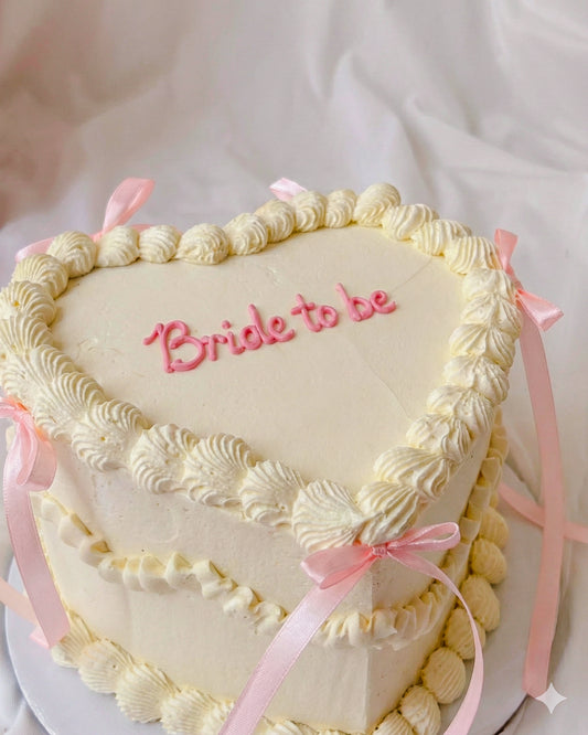 Heart-shaped cake with 'Bride to be' text and pink ribbons on a white background