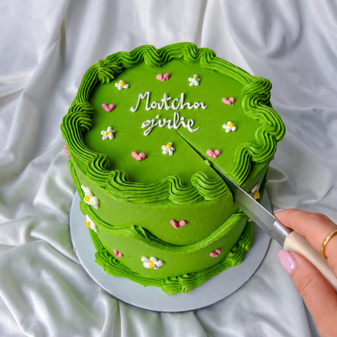 Green cake with 'Matcha girlie' text being sliced on a white fabric background