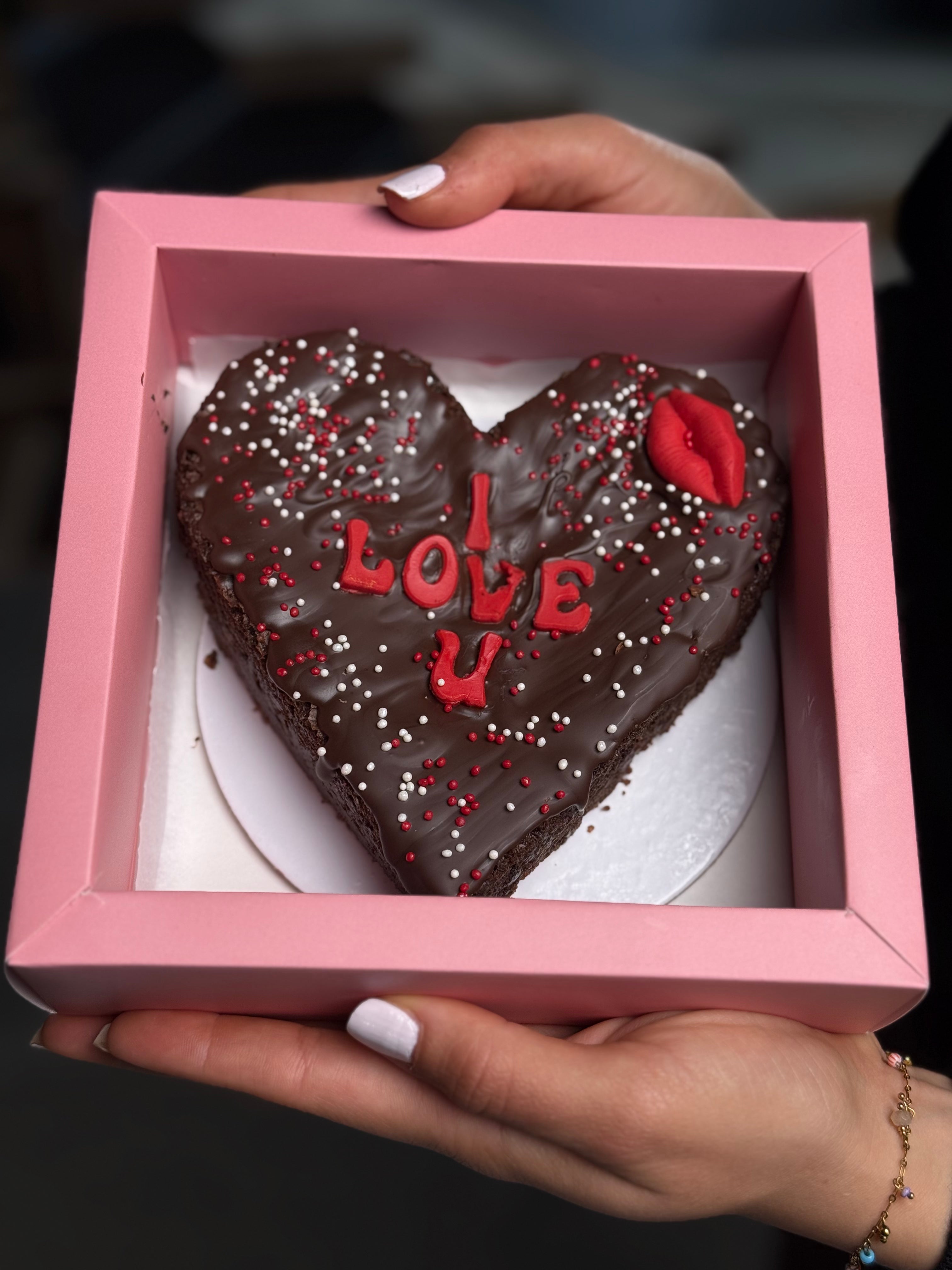 Heart-shaped chocolate cake with 'I LOVE U' chocolate text in a pink box held by a person.