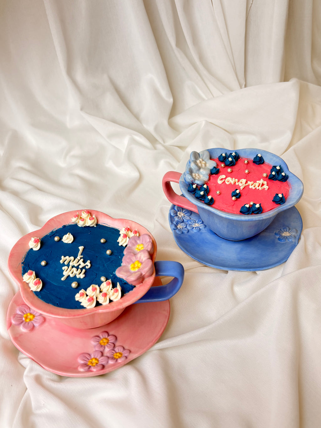 Two decorative teacups with messages 'I miss you' and 'Congratulations' on a white fabric background.