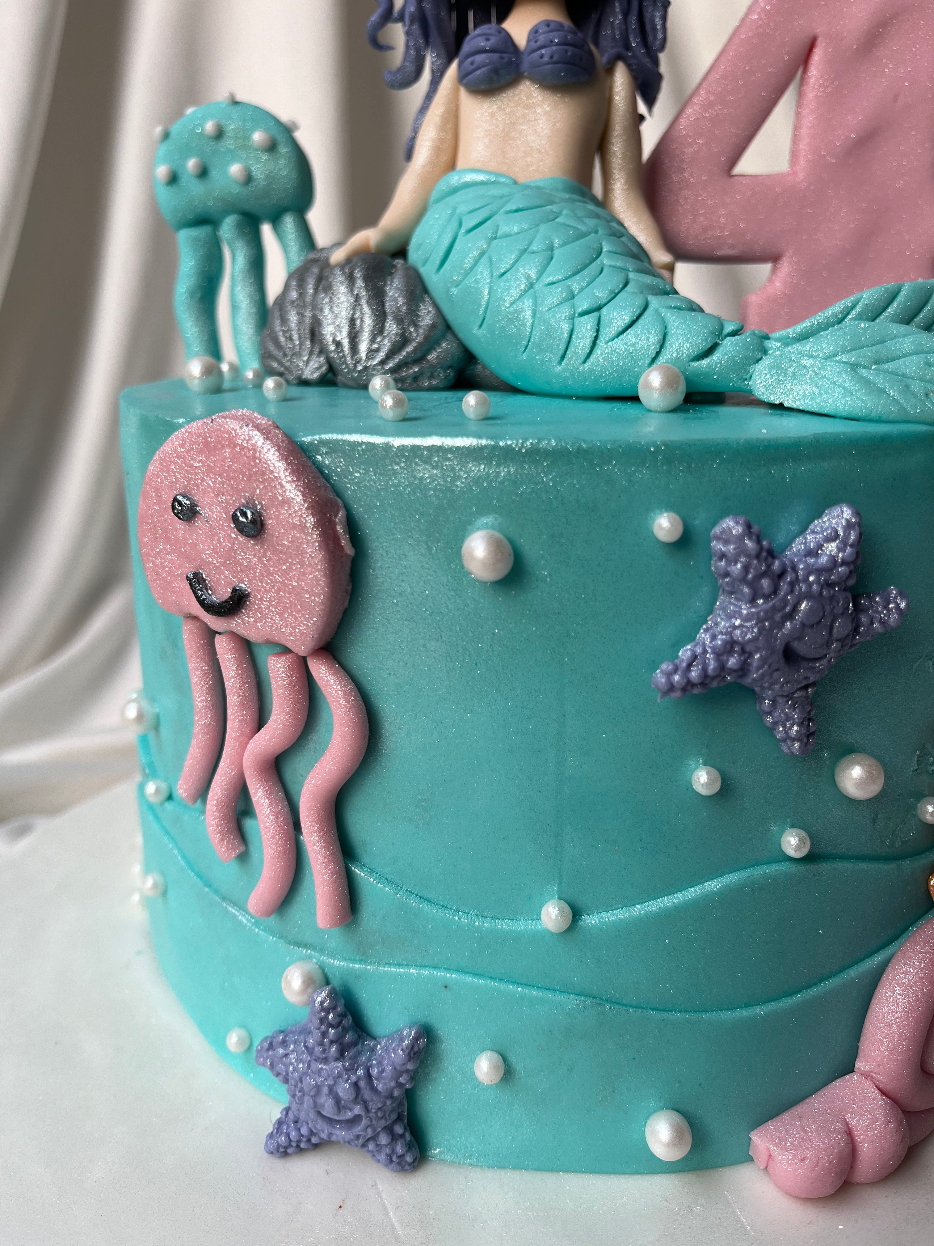 Teal cake with mermaid and sea creature decorations on a white background