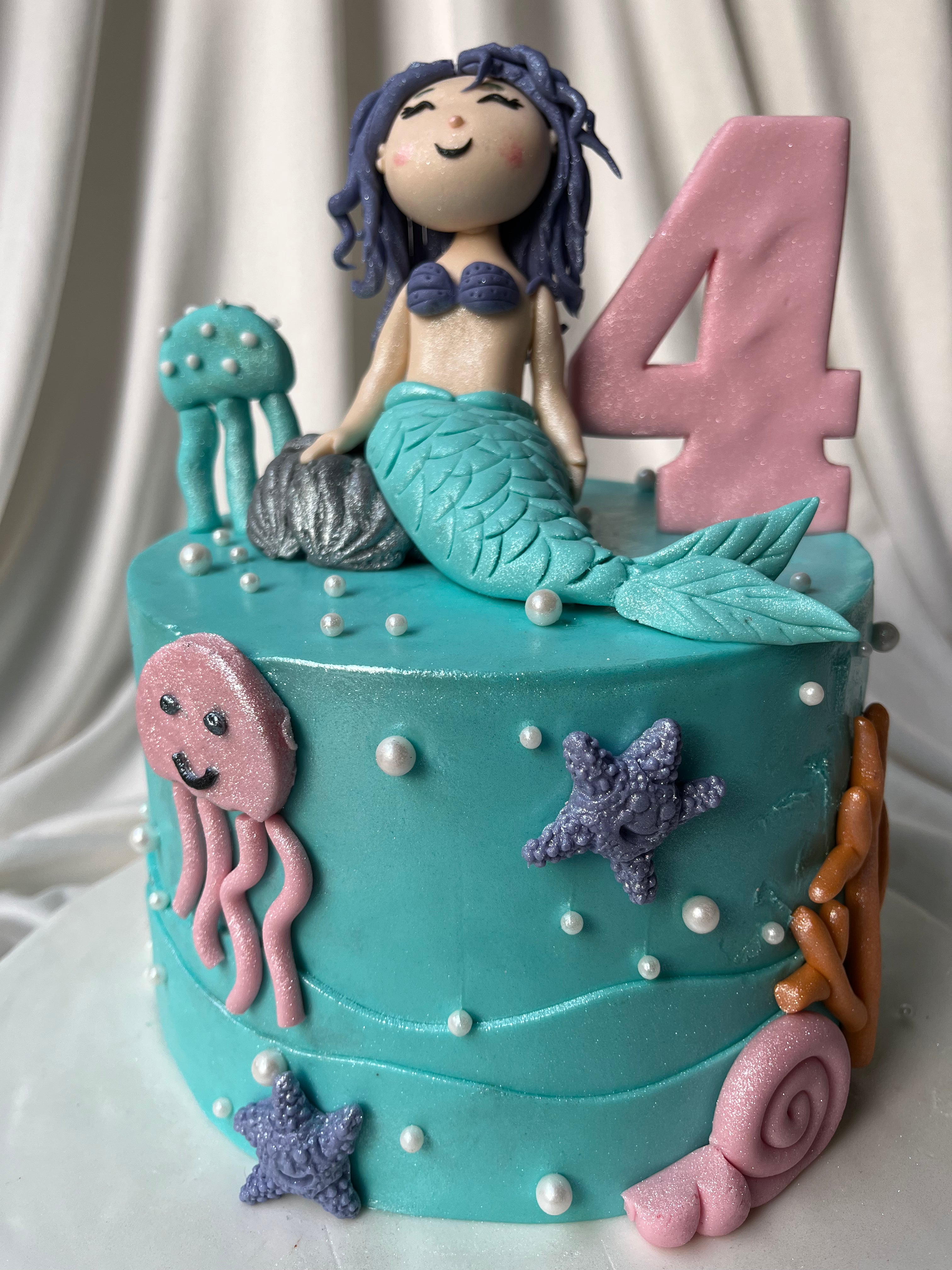 Cake with mermaid figurine, number 4, jellyfish, and starfish decorations on a white background.