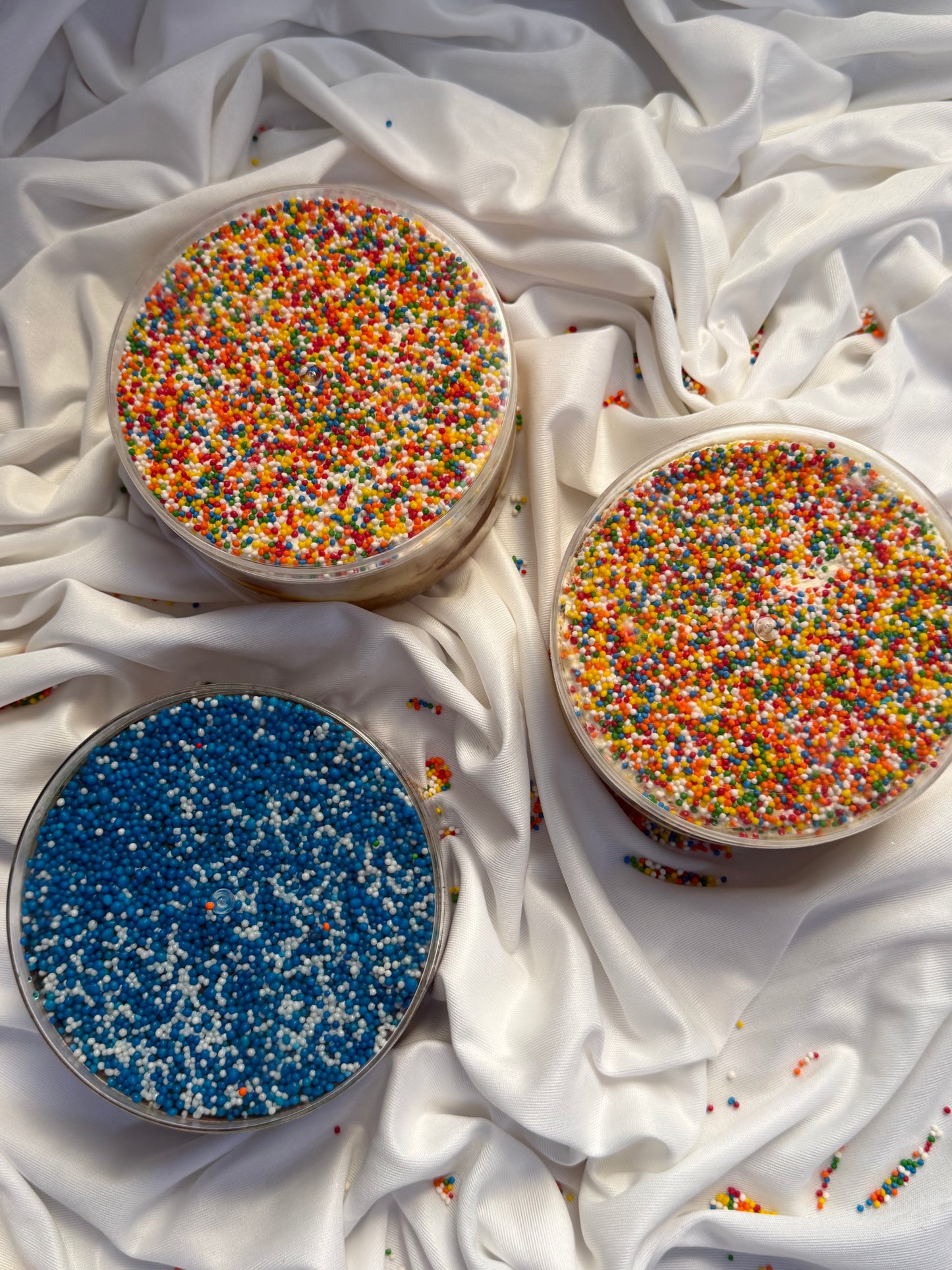 Three round containers filled with colorful sprinkles on a white fabric background