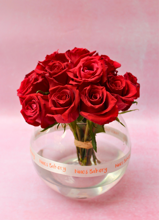 A round glass vase containing a bouquet of 20 red roses, displayed on a pink background.