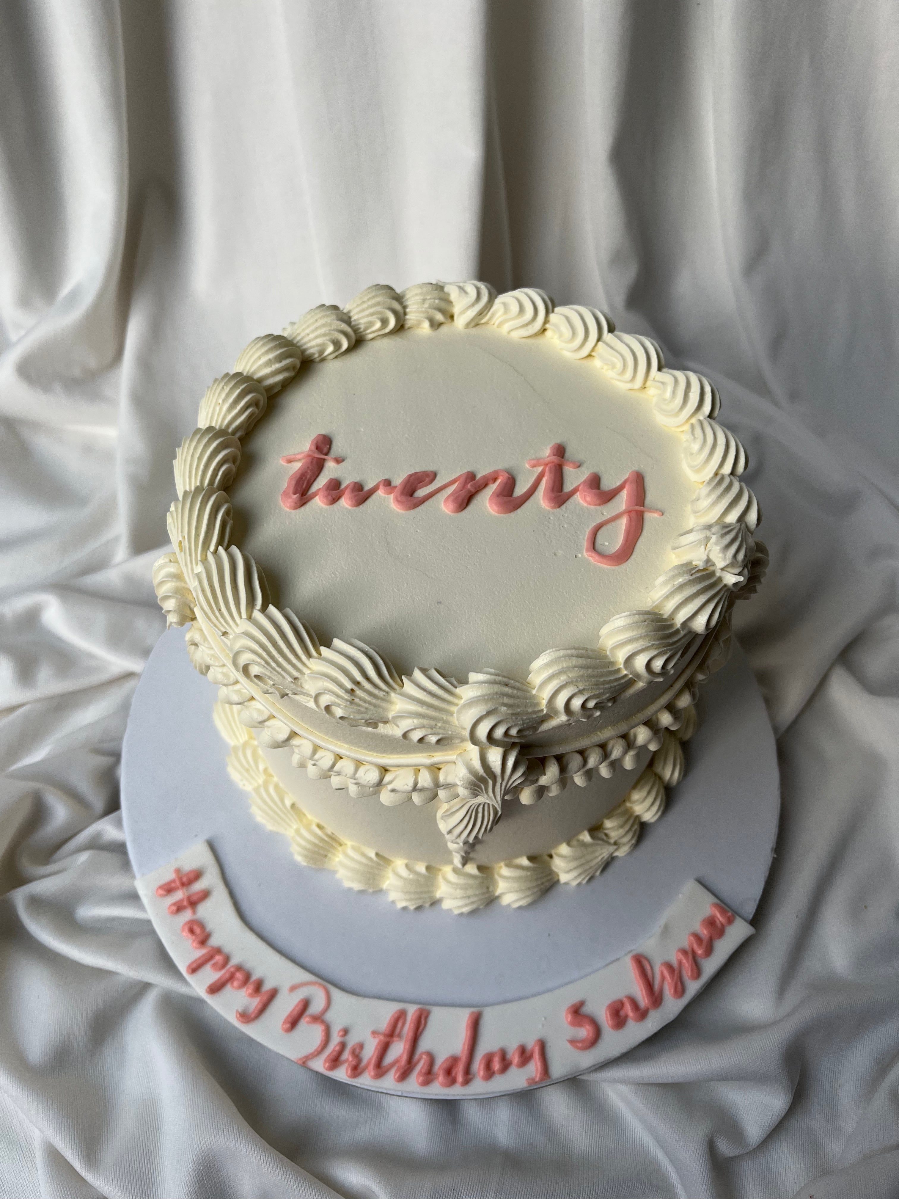 Two-tier birthday cake with 'twenty' and 'Happy Birthday Salva' text on a white fabric background