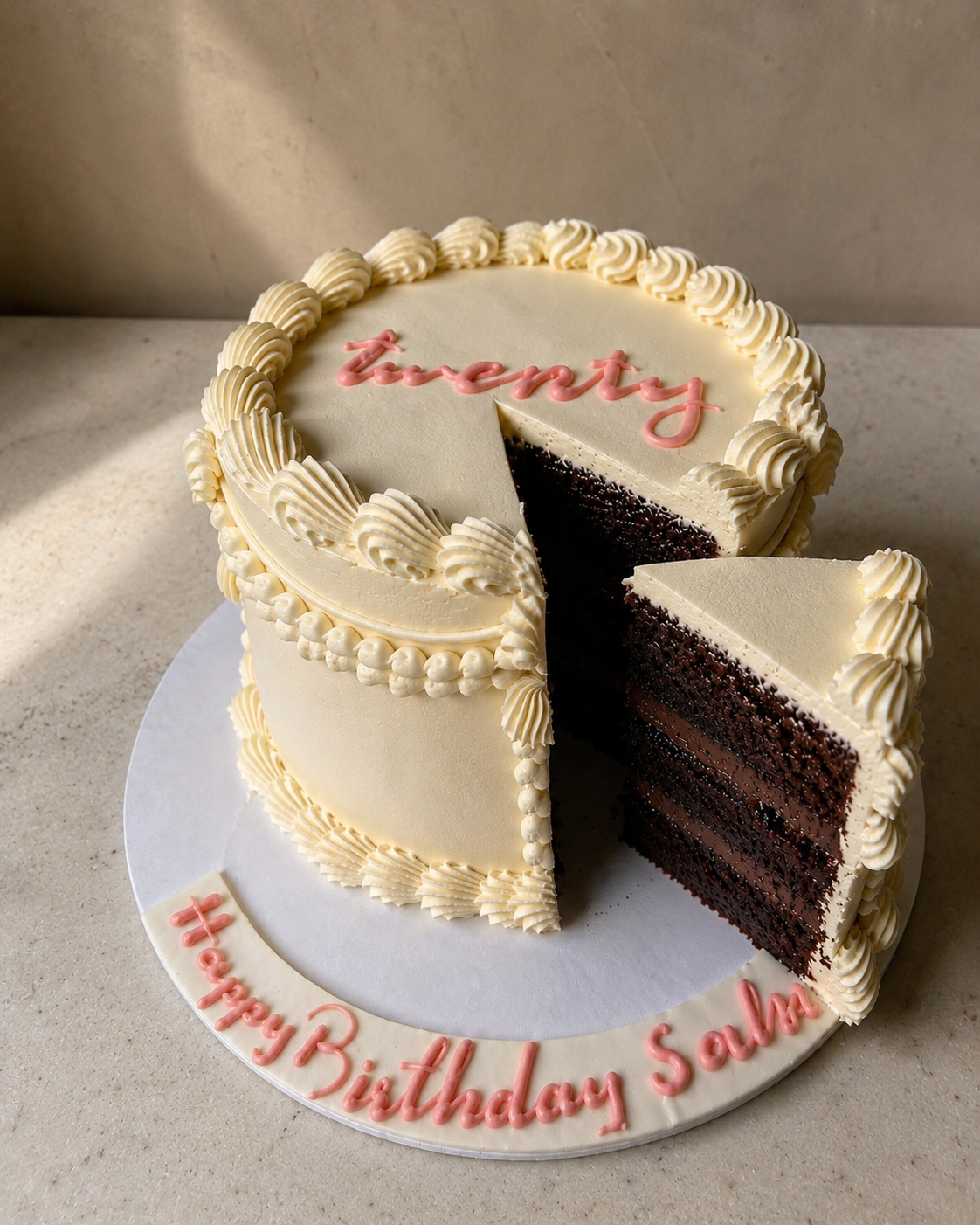 Chocolate cake with white frosting and 'twenty' written on it, with a slice taken out, on a light surface.