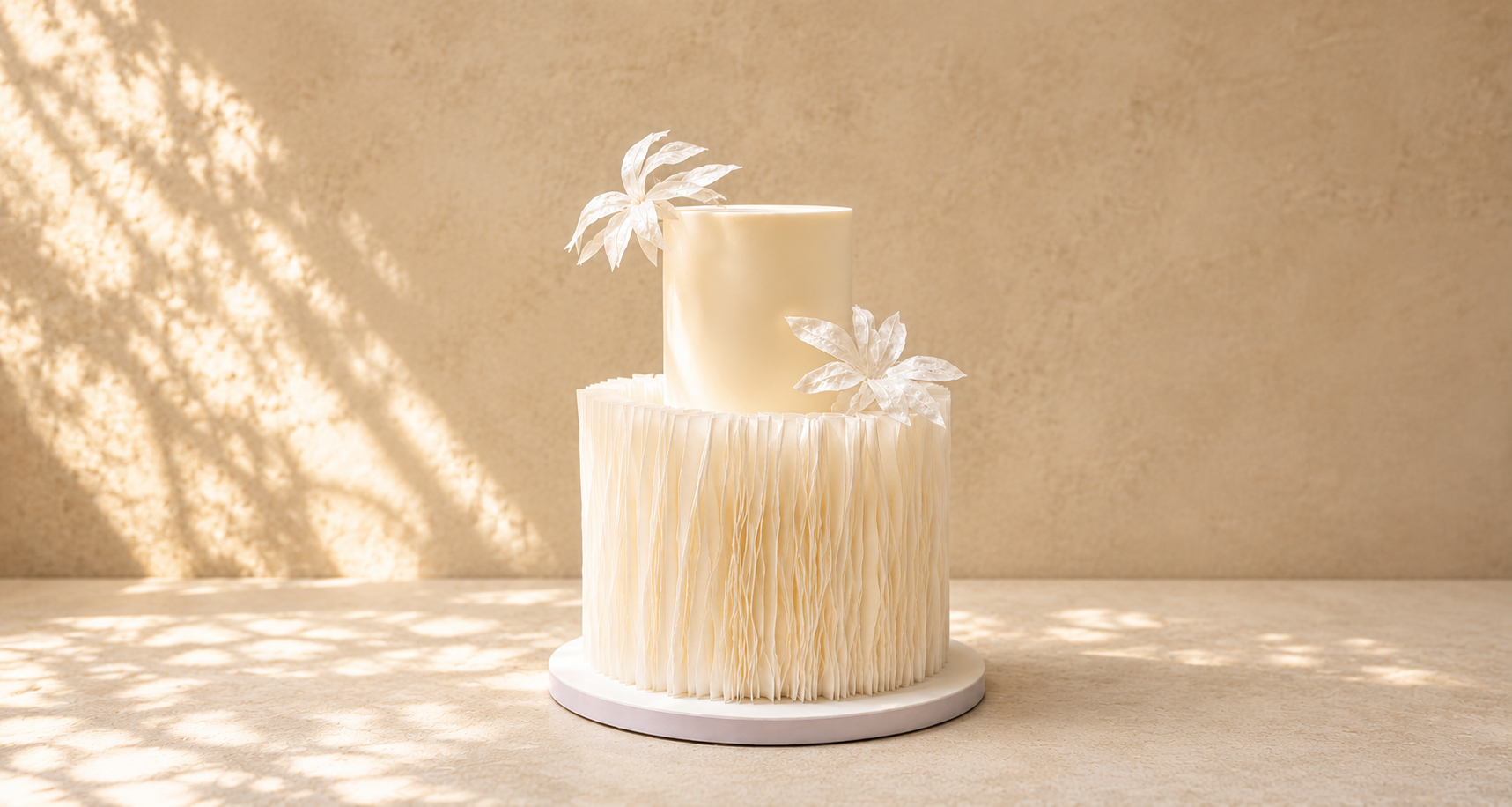 A two-tier cream-colored cake with delicate white floral decorations, displayed on a round white base against a textured concrete wall with natural leaf shadows.